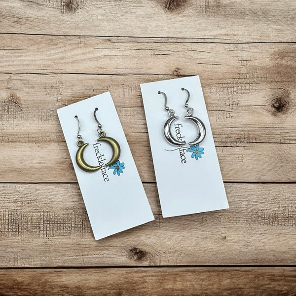 Bronze and silver crescent hoop earrings displayed on Freckle Face Jewellery cards over a rustic wood background.