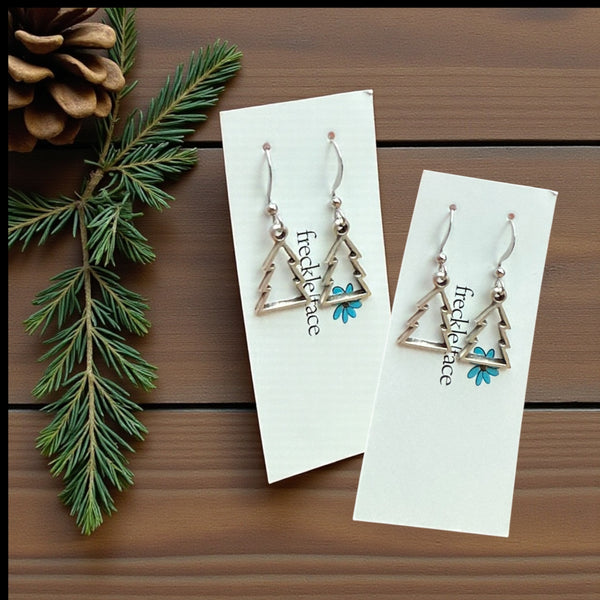 Pair of cookie cutter christmas tree earrings on Freckleface branded cards with a pine cone and branch on a wooden surface.