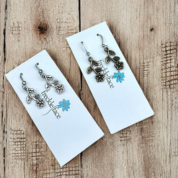 Two pairs of floral vine earrings in antique silver and bronze displayed on Freckle Face earring cards over a rustic wood background.