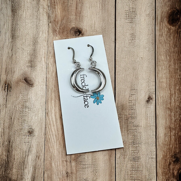 Shiny silver crescent earrings on white Freckle Face Jewellery card, styled against natural wood grain.