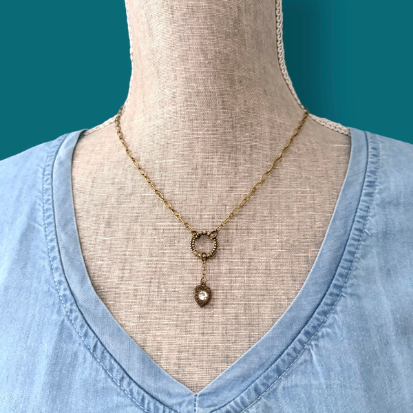 Heartstring necklace in antique brass worn on a mannequin, featuring a textured circle connector and a delicate heart charm centered on the chest.