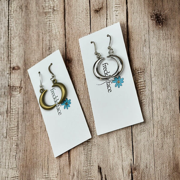 Crescent hoop earrings in bronze and silver tones displayed side-by-side on Freckle Face Jewellery cards, rustic wood surface.