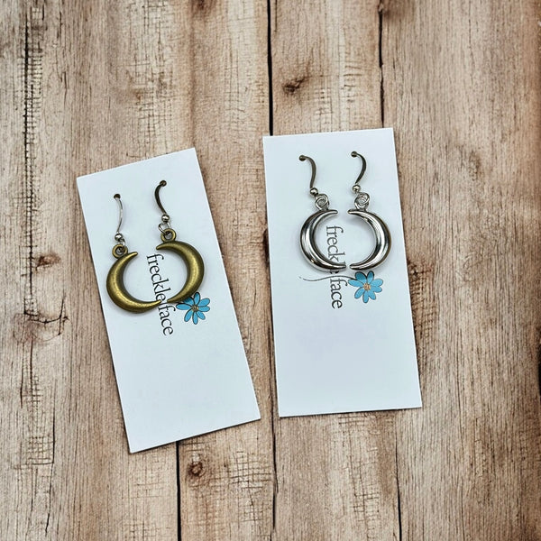 Minimalist crescent hoop earrings in silver and bronze finishes on white Freckle Face Jewellery cards, flat lay on wood.