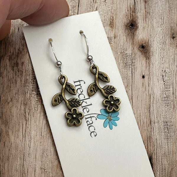 Hand holding Freckle Face card with boho bronze wildflower earrings on rustic wood background.