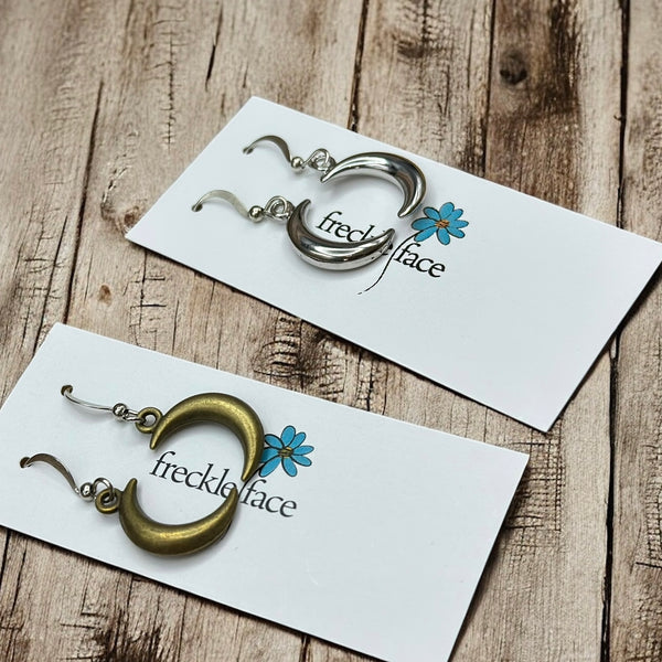 Close-up angled view of silver and bronze crescent hoop earrings on branded earring cards, placed on a wood surface.