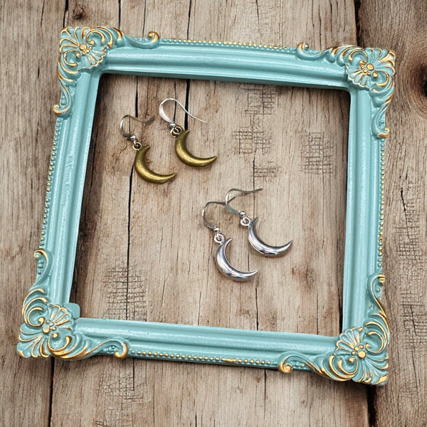 Bronze and silver crescent moon earrings displayed inside a teal vintage frame on rustic wood background.