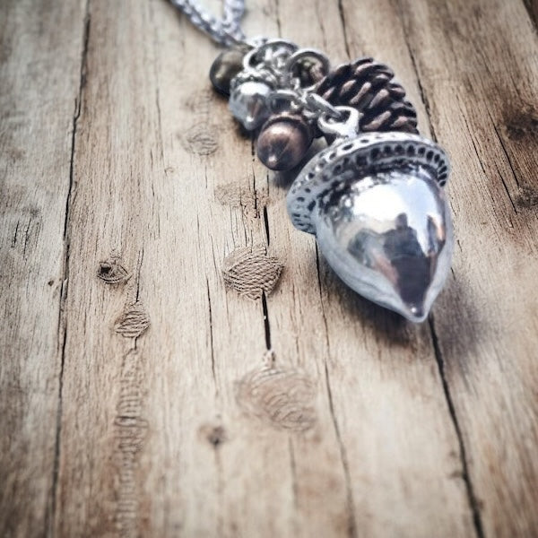 Close-up of a silver acorn pendant with pinecone and acorn charms on a rustic wooden surface.