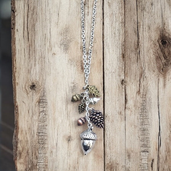 Hanging woodland charm necklace with silver acorn, pinecone, and leaf charms on a natural wood background.