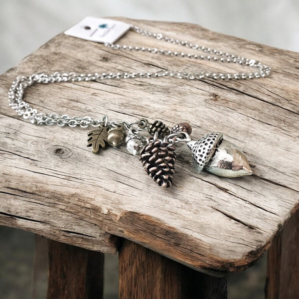 Forest-inspired charm necklace with acorn, pinecone, leaf, and nut charms displayed on a rustic wood table.