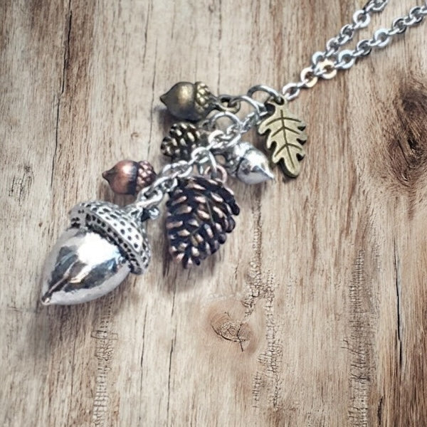 Woodland charm necklace hanging against weathered wood, featuring acorn, pinecone, leaf, and nut charms.