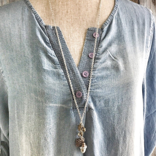Long woodland-themed charm necklace styled on a denim blouse, featuring acorn and forest charms.