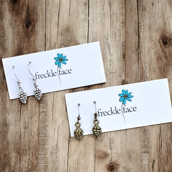 Two pairs of pinecone-shaped earrings displayed on white cards with 'freckle face' branding. One pair is silver, and the other is brass, both with intricate textured scales.