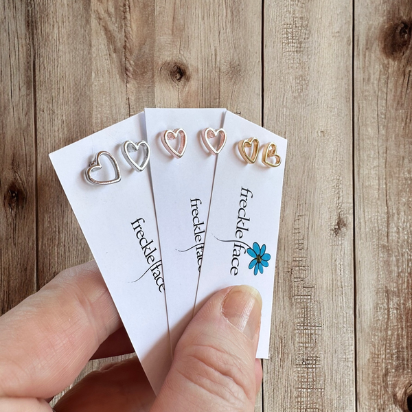 Set of minimalist tiny open heart studs in silver, gold, and rose gold displayed in hand