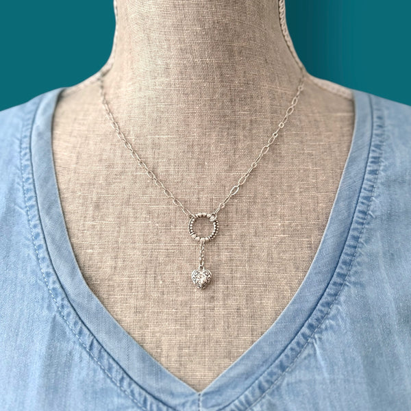 Close-up of the Heartstring necklace in antique silver, highlighting the detailed circular link and heart charm against a soft blue top.