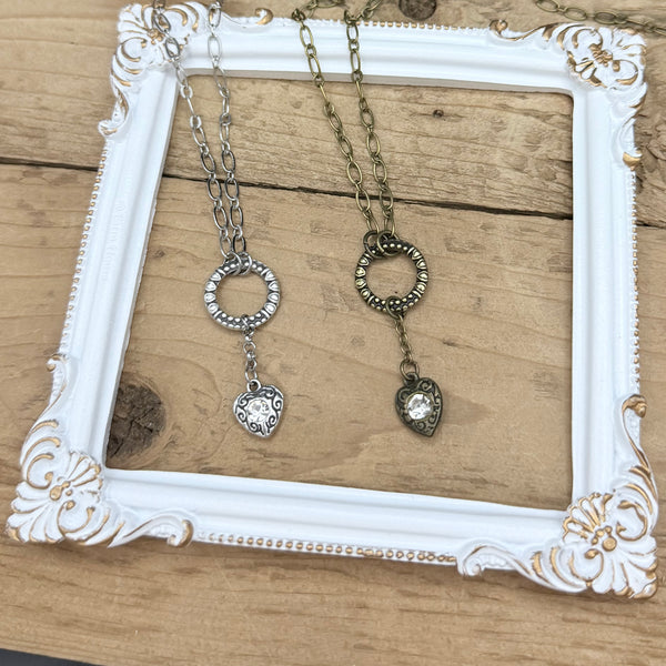 Flat lay of Heartstring necklaces in antique silver and antique brass, showing oval link chains and heart drop charms on a natural wood background.
