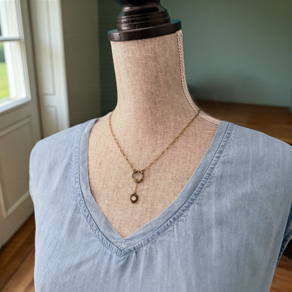 Heartstring necklace in antique brass worn on a mannequin, featuring a textured circle connector and a delicate heart charm centered on the chest.
