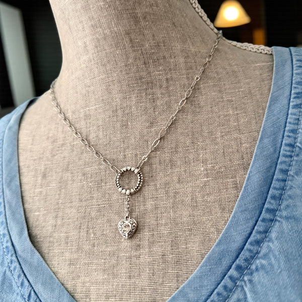 Close-up of the Heartstring necklace in antique silver, highlighting the detailed circular link and heart charm against a soft blue top.